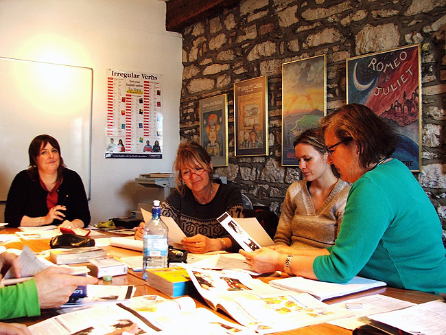 Adults at a table during a lesson surrounded by open english language textboots in a cosy classroom