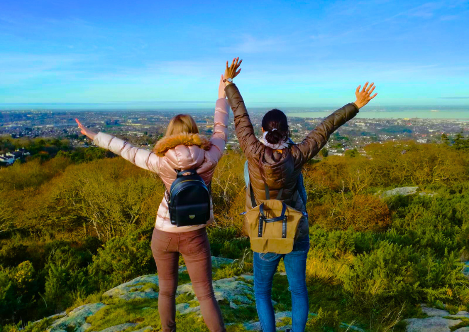 Two students with their arms in the air staring out over a mountain view.